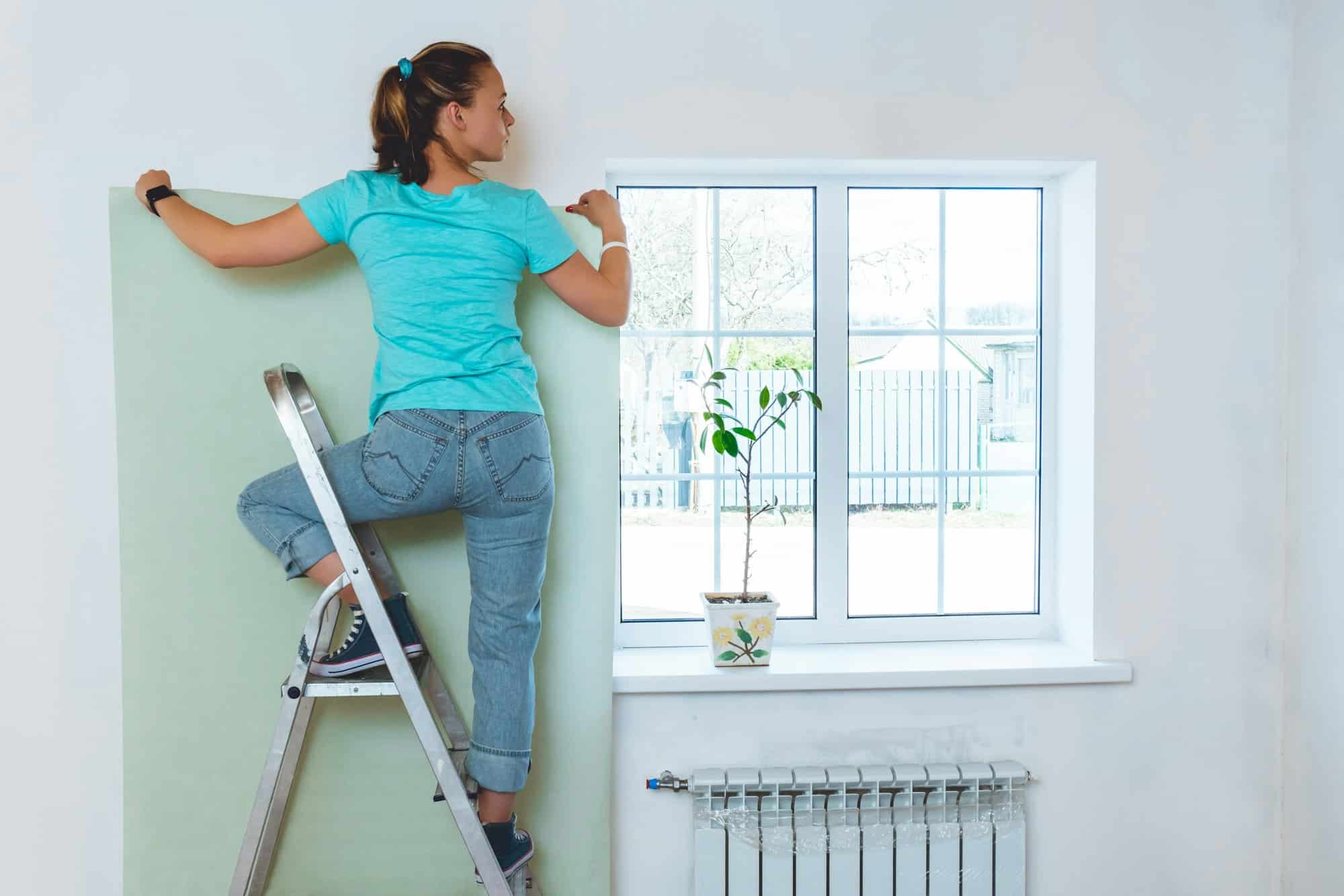 Woman in jeans and turquoise T-shirt standing on stepladder and holding piece of wallpaper